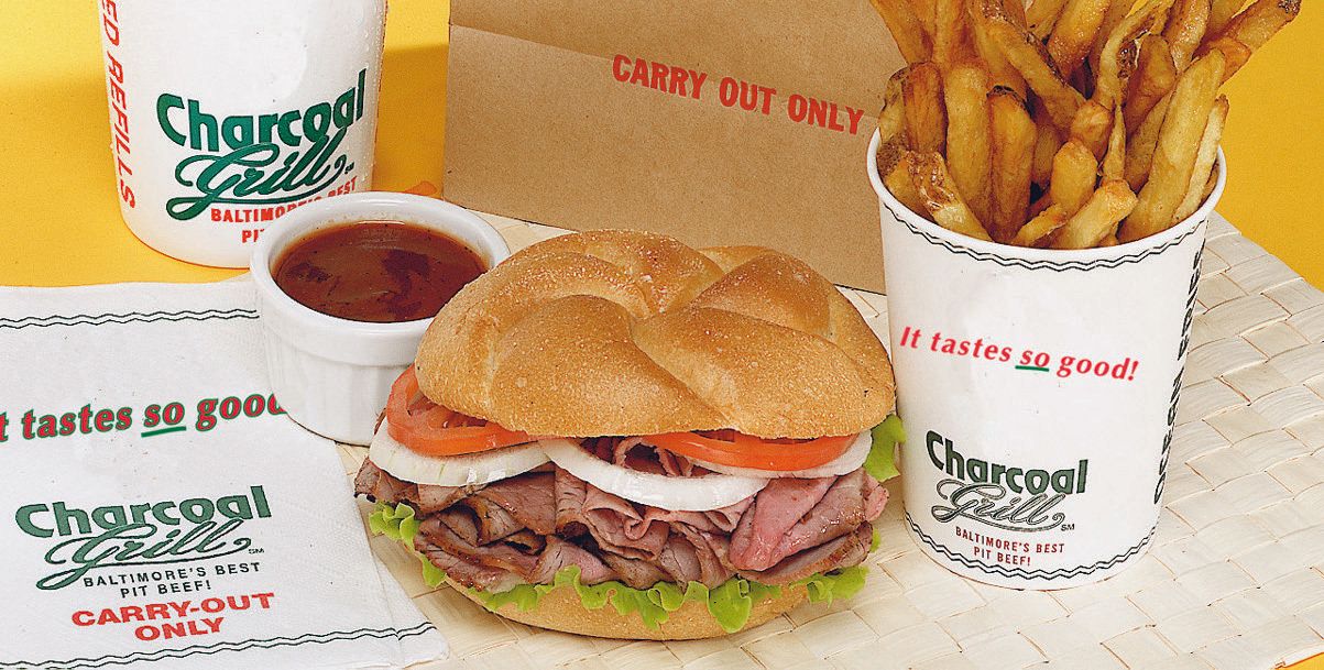 Pit beef sandwich with tomato and onion beside a cup of fresh-cut fries and barbecue sauce